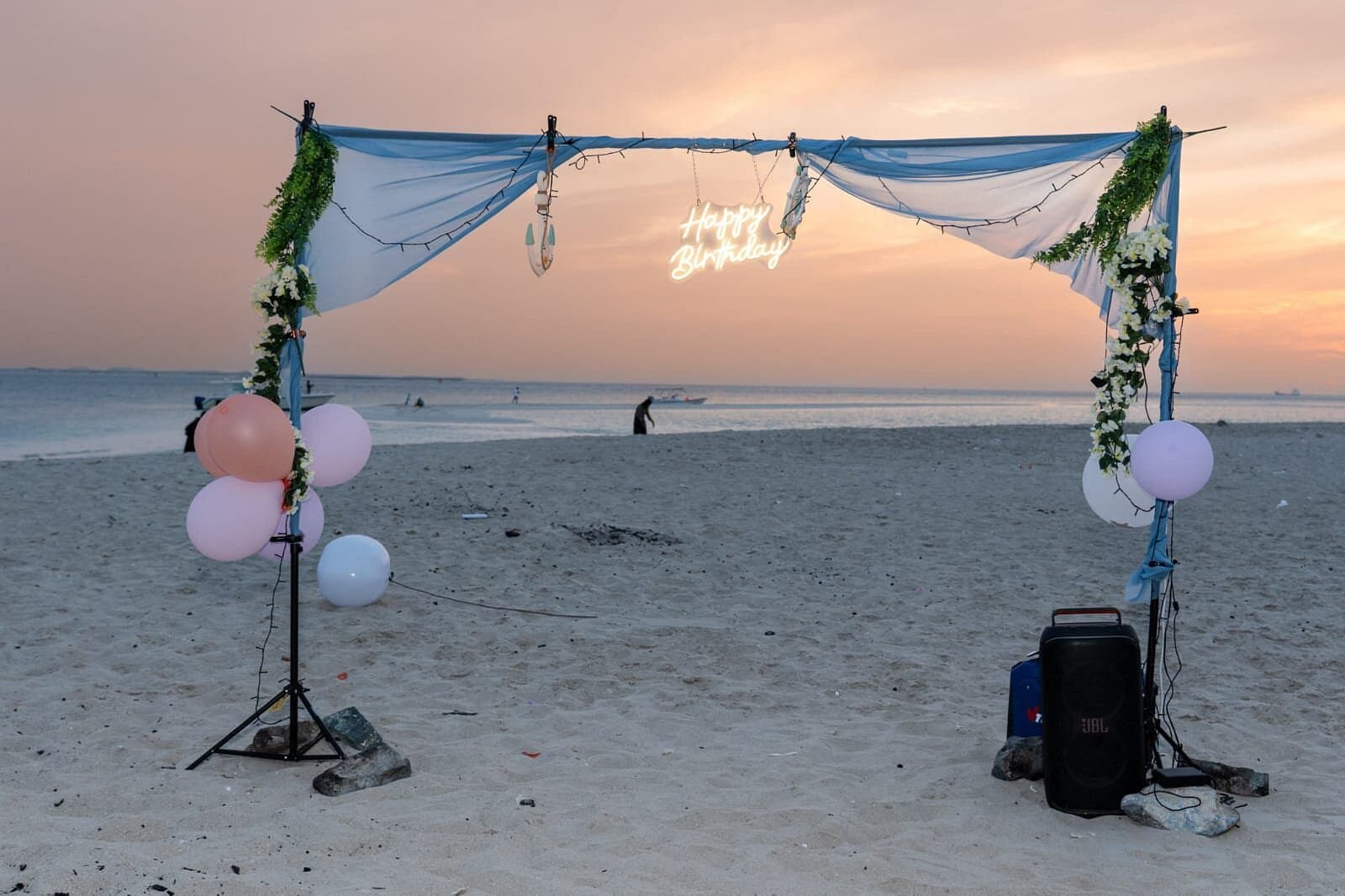 Happy Birthday neon sign under a draped beach arch with balloons at sunset